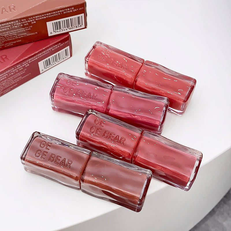 Four lip glosses with 'GE BEAR' branding on a white surface, with two boxes in the background.