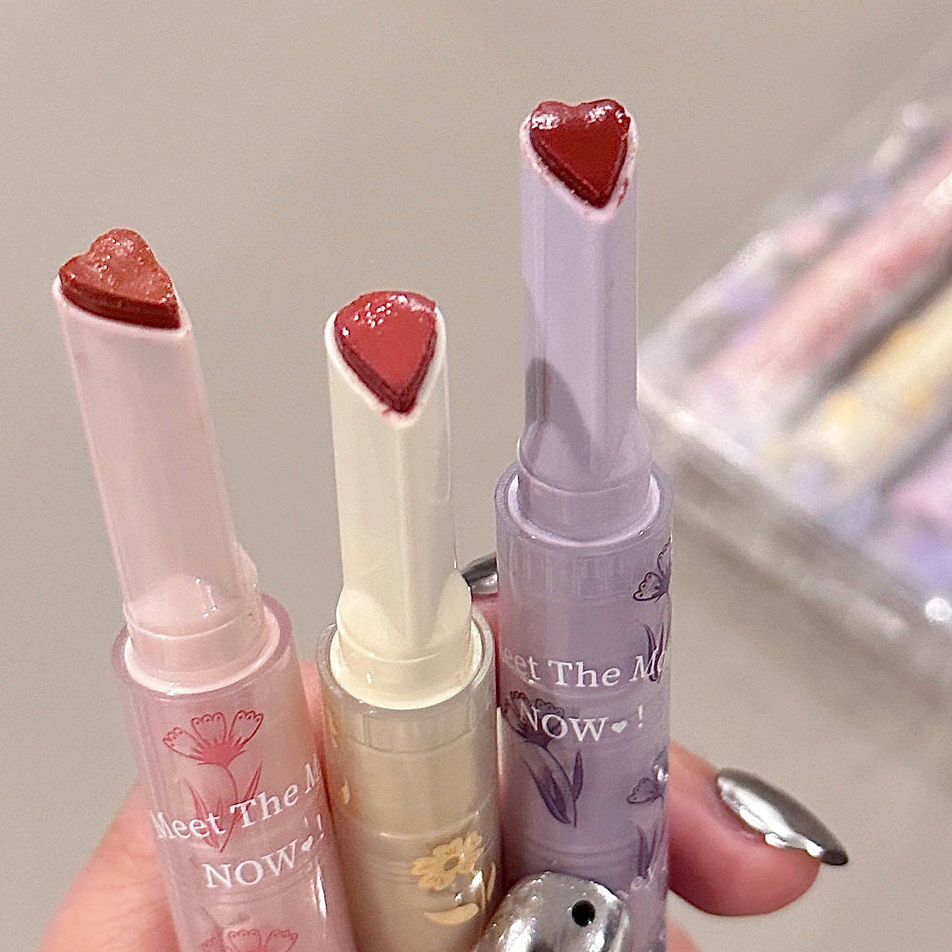 Three lip glosses with heart-shaped caps held by a hand against a neutral background.