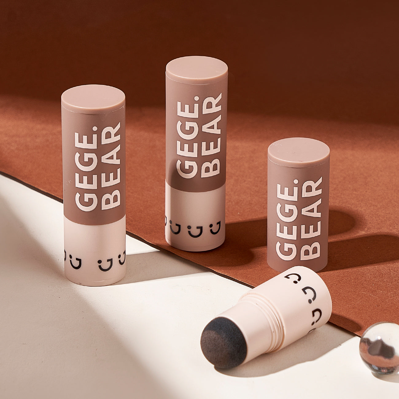 Three GEGE BEAR cosmetic tubes on a brown surface with a reflective white surface underneath.