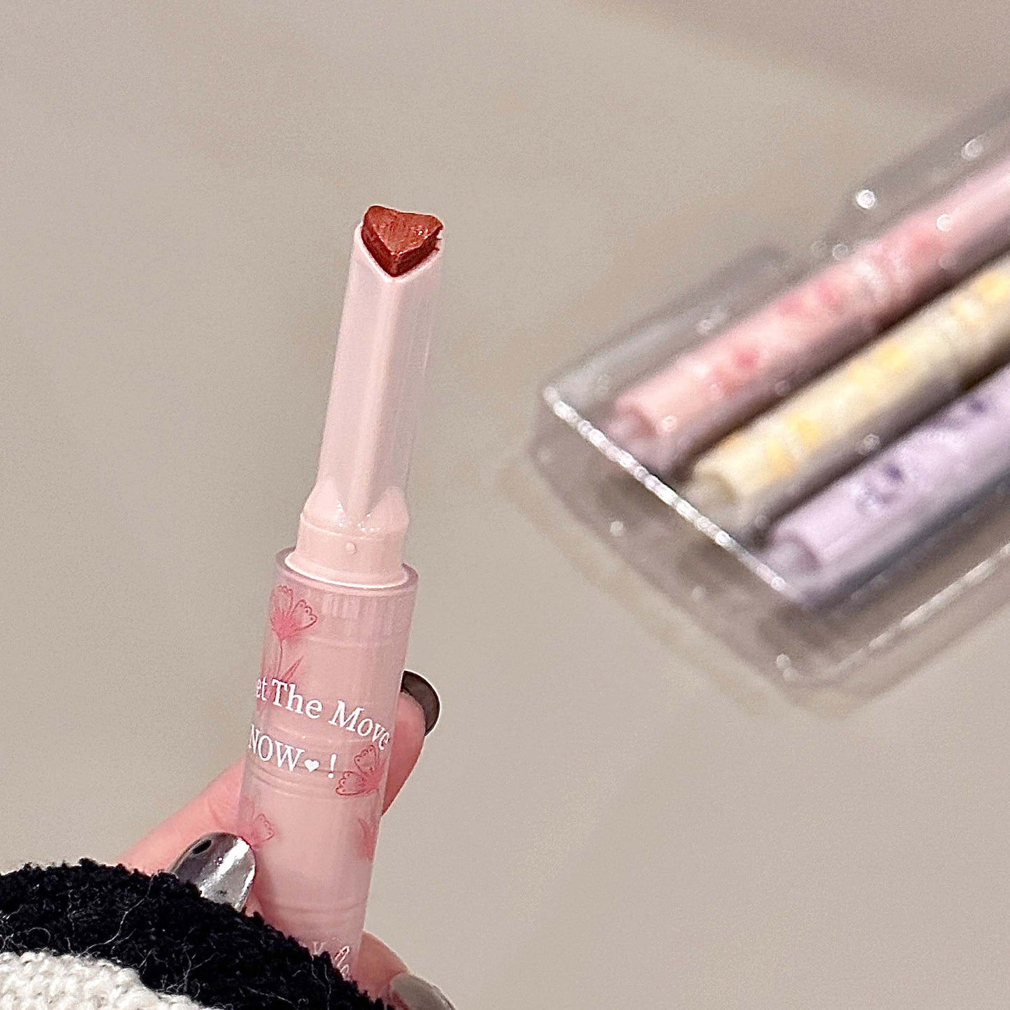 Pink lip gloss with heart-shaped cap held by a hand, with more lip glosses in the background.