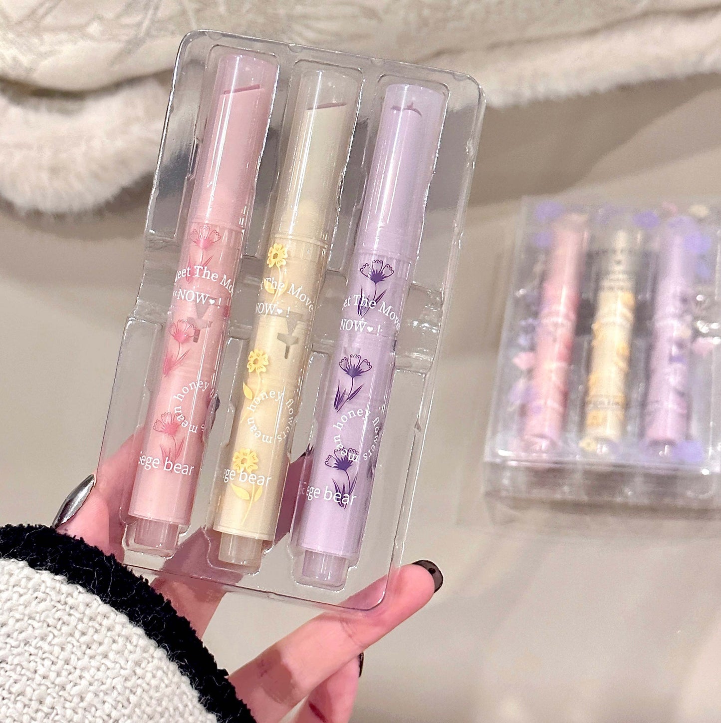 Set of three lip glosses in clear packaging held by a person.