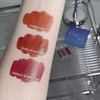 Lipstick swatches on a person's arm with product names and codes, against a blurred background.